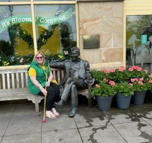 Amanda on a bench with her long yellow cane, she is sitting next a statue of a seated man