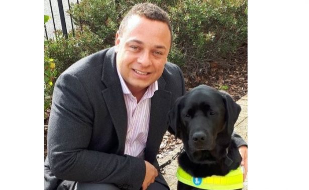 Dan smiling, wearing a suit and kneeling down with his arm around his guide dog Zodiac, a black, labrador.