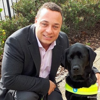 Dan smiling, wearing a suit and kneeling down with his arm around his guide dog Zodiac, a black, labrador.