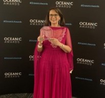 Shahina wearing an elegant, long, red Asian dress and scarf, holding her award.