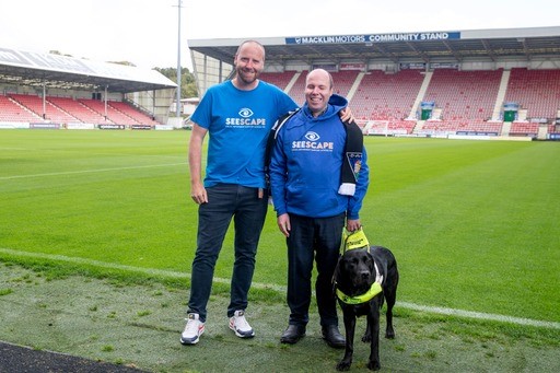 Graham, Stuart and his guide dog standing by the side of the football pitch at Dunfermline Athletic Football Club.