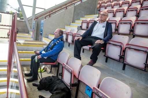 Stuart sitting in a stadium at Dunfermline Athletic Football Club with his guide dog lying at his feet. Dave Henderson is sitting behind them providing audio commentary.