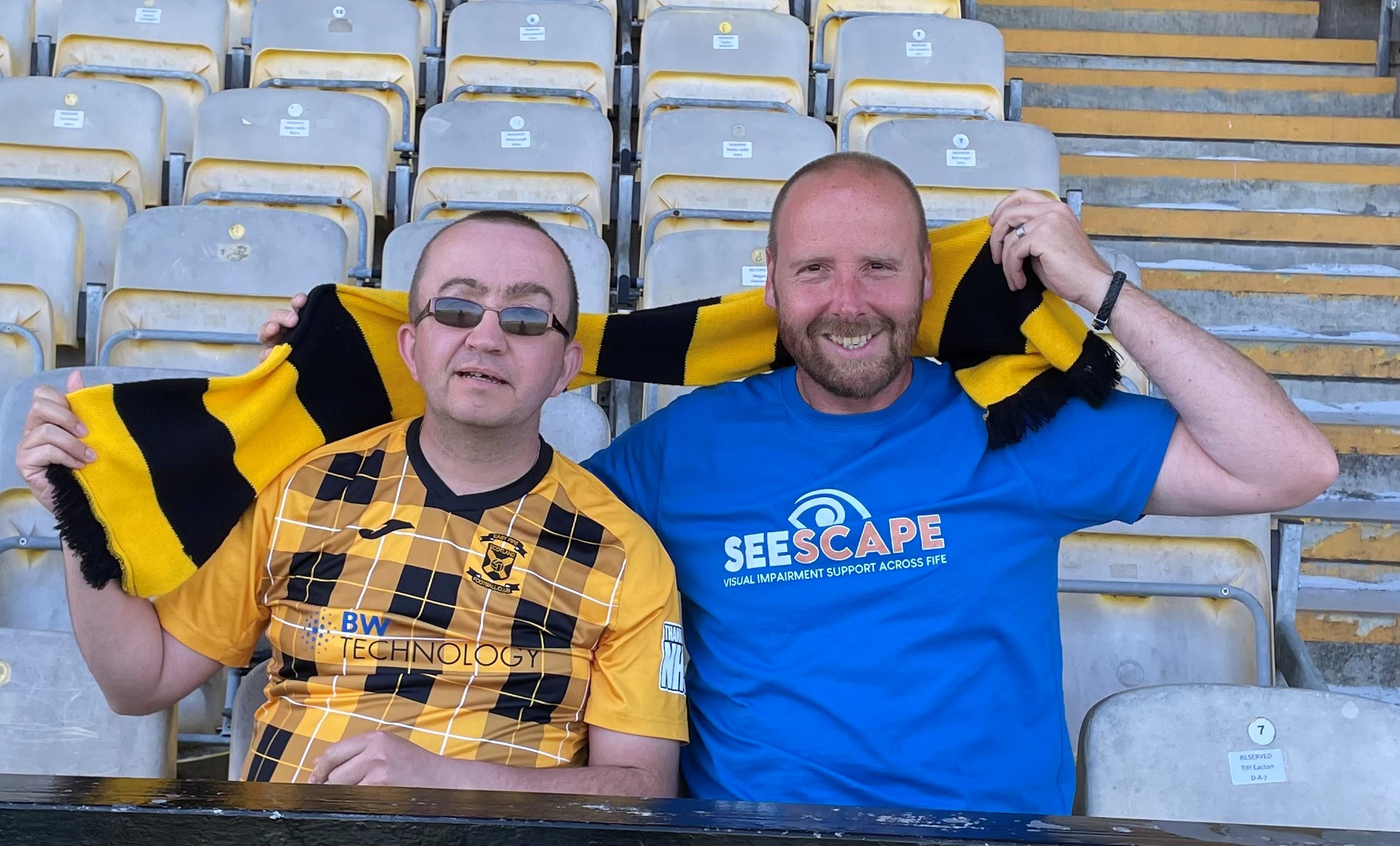 Sandy Allan and Graham Ross sitting in the stands at East Fife Football Club holding the club's yellow and black scarf behind them.