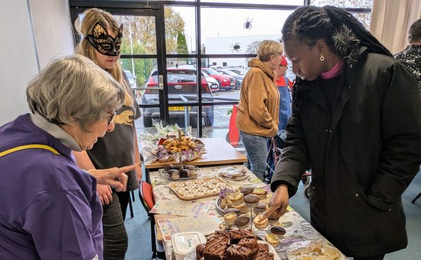 Two women (one wearing a cat face mask, serving another woman looking at a table full of cakes.