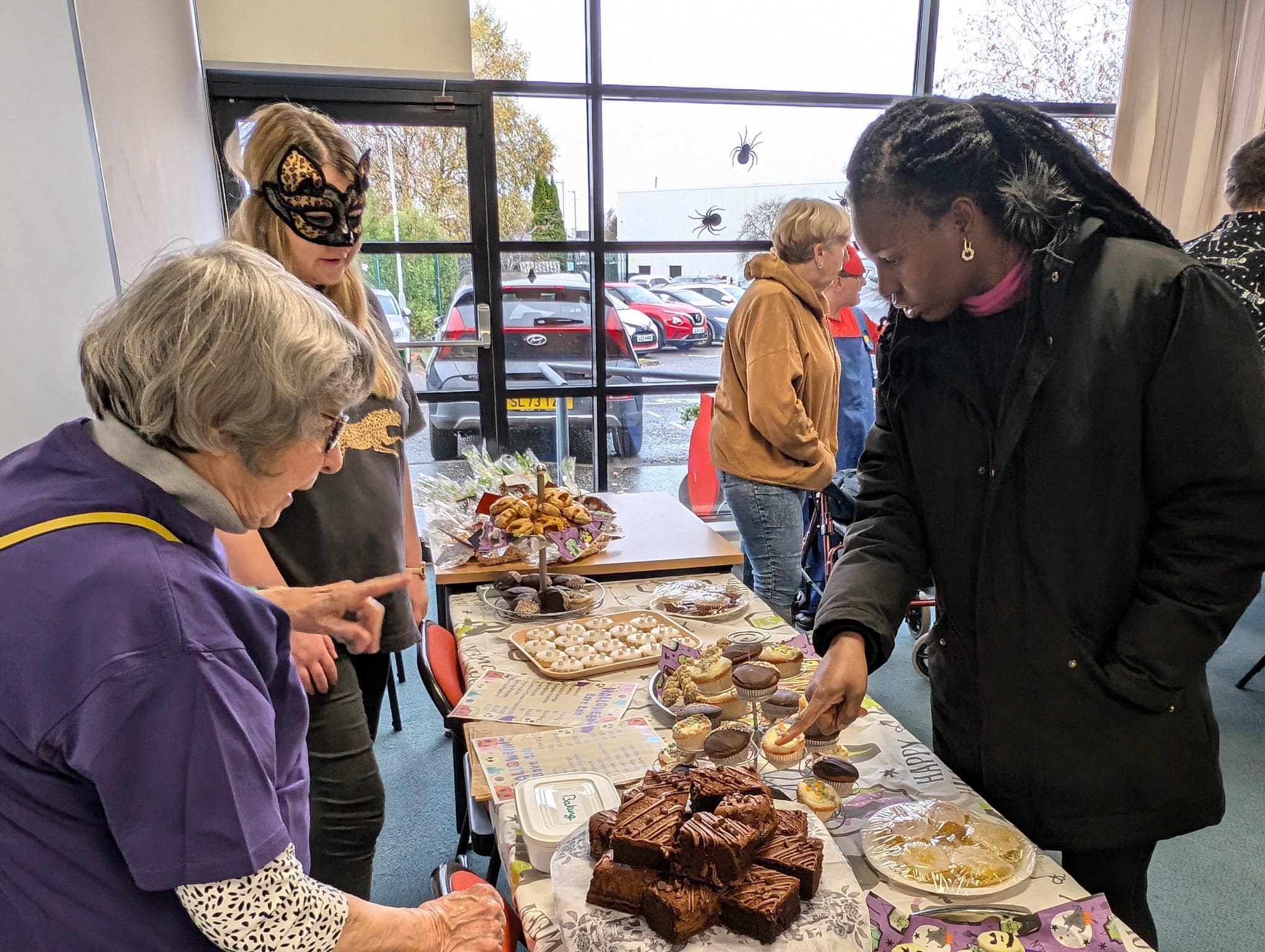 Two women (one wearing a cat face mask, serving another woman looking at a table full of cakes. 