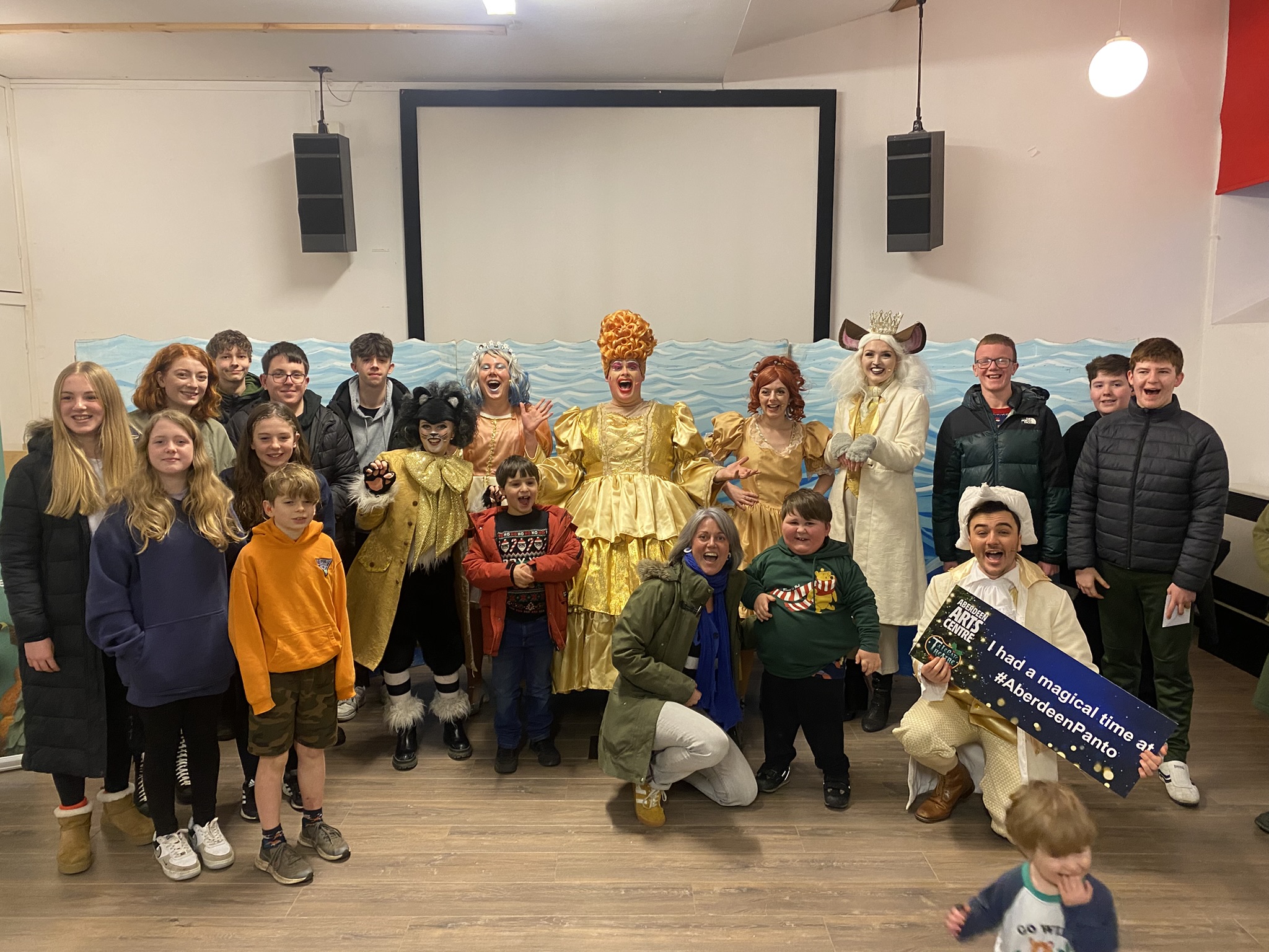 Cast of Dick Whittington at Aberdeen Arts Centre in full costume with children and young people and NESS team. Dick Whittington is kneeling and holding a sign that says "I had a magical time at #AberdeenPanto." and has "Aberdeen Arts Centre" on the left hand of the sign. Photo by NESS.