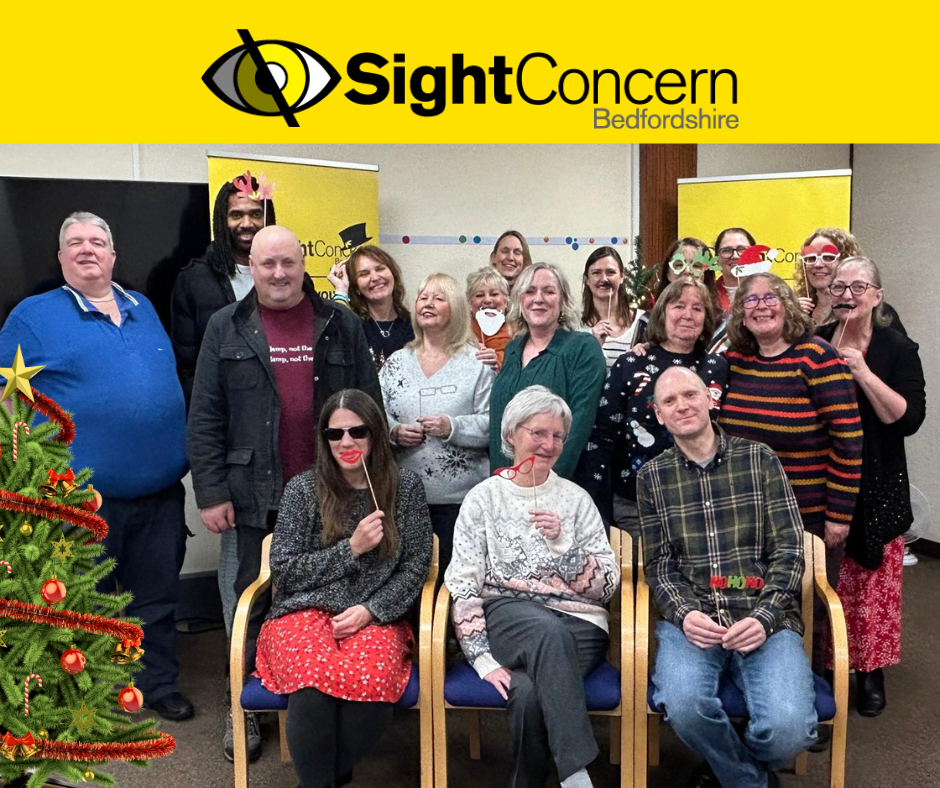 Sight Concern Bedfordshire team and service users holding sticks with glasses, santa beard, santa hats and the words "ho, ho, ho," There is a decorated christmas tree on the left hand side. The Sight Concern Bedfordshire logo is at the top.