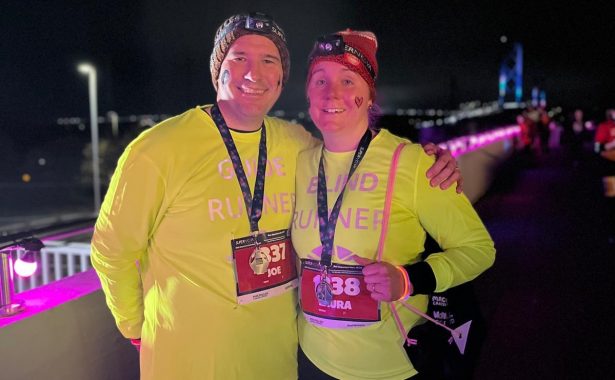 Joe with his arm around Laura standing outdoors at night, wearing yellow sweatshirts that say "Blind Runner" and wearing medals around their necks.