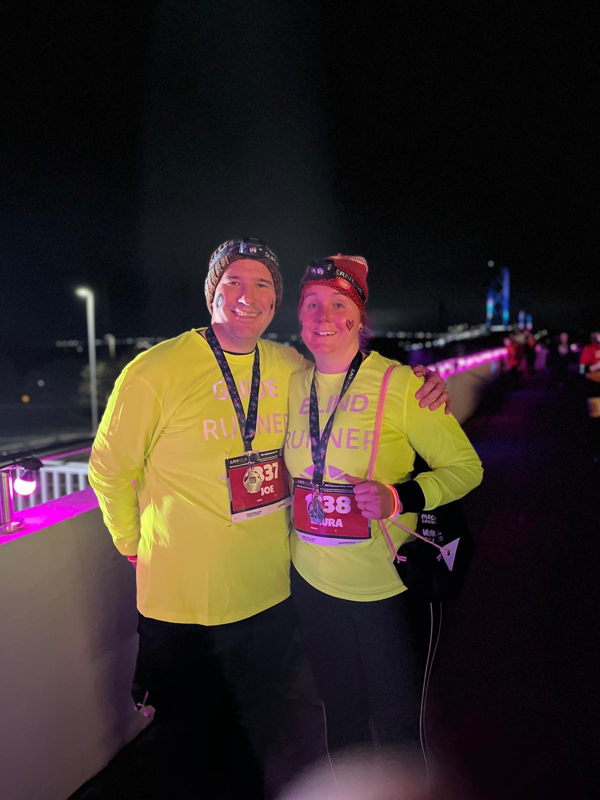 Joe with his arm around Laura standing on the Forth Bridge at night, wearing yellow sweatshirts that say "Blind Runner" and wearing medals around their necks.