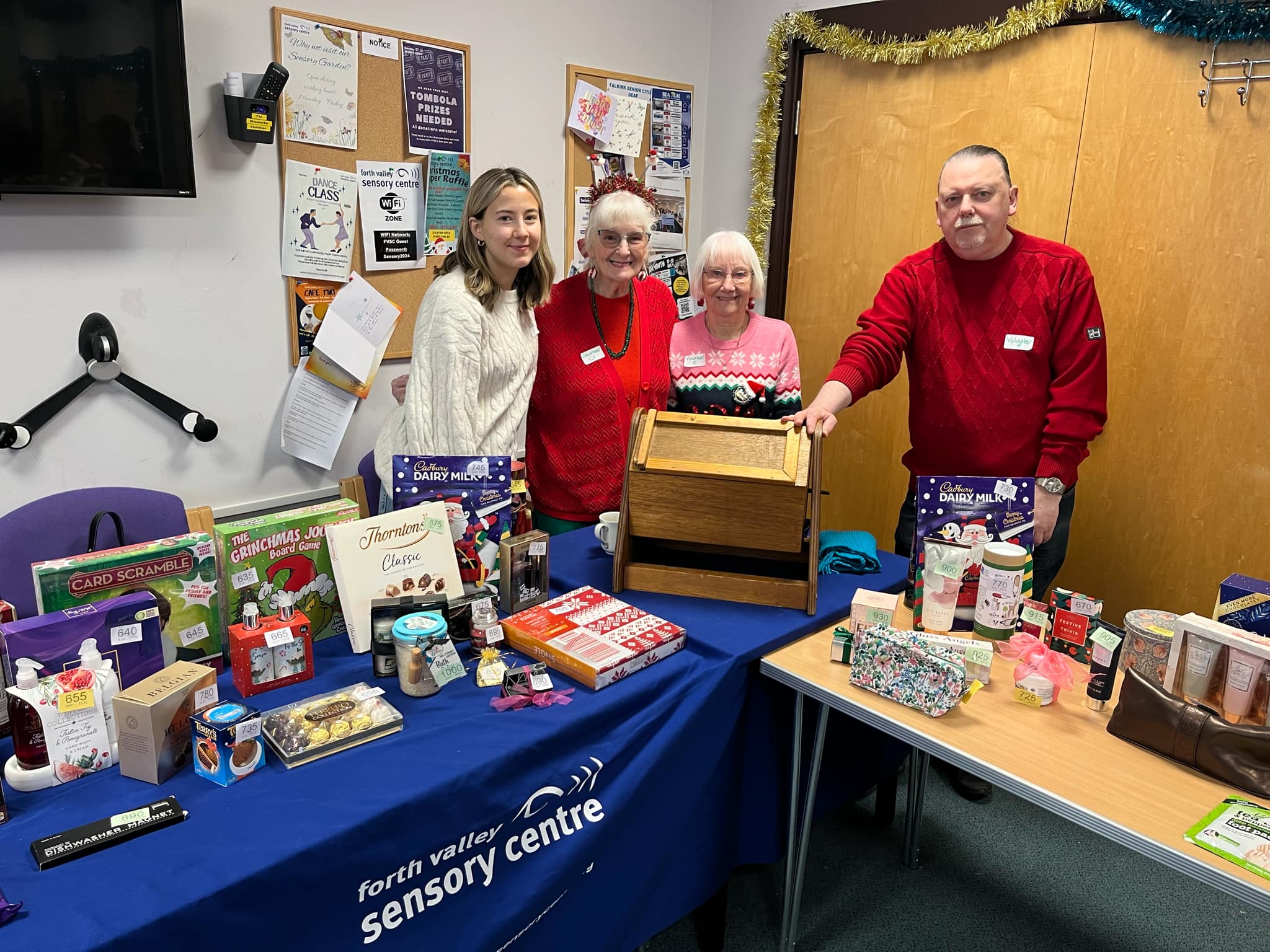 Four people behind a tomobola with various prizes on tables including chocolates, toiletries and games.