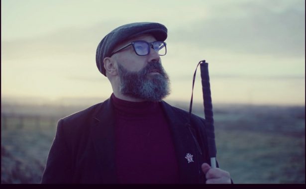 Dave Steele standing outdoors with hazy background of a field and cloudy sky. Dave is holding up his cane. He is wearing a grey cap, glasses, dark blazer with a motif on the lapel and a maroon turtle neck sweater.