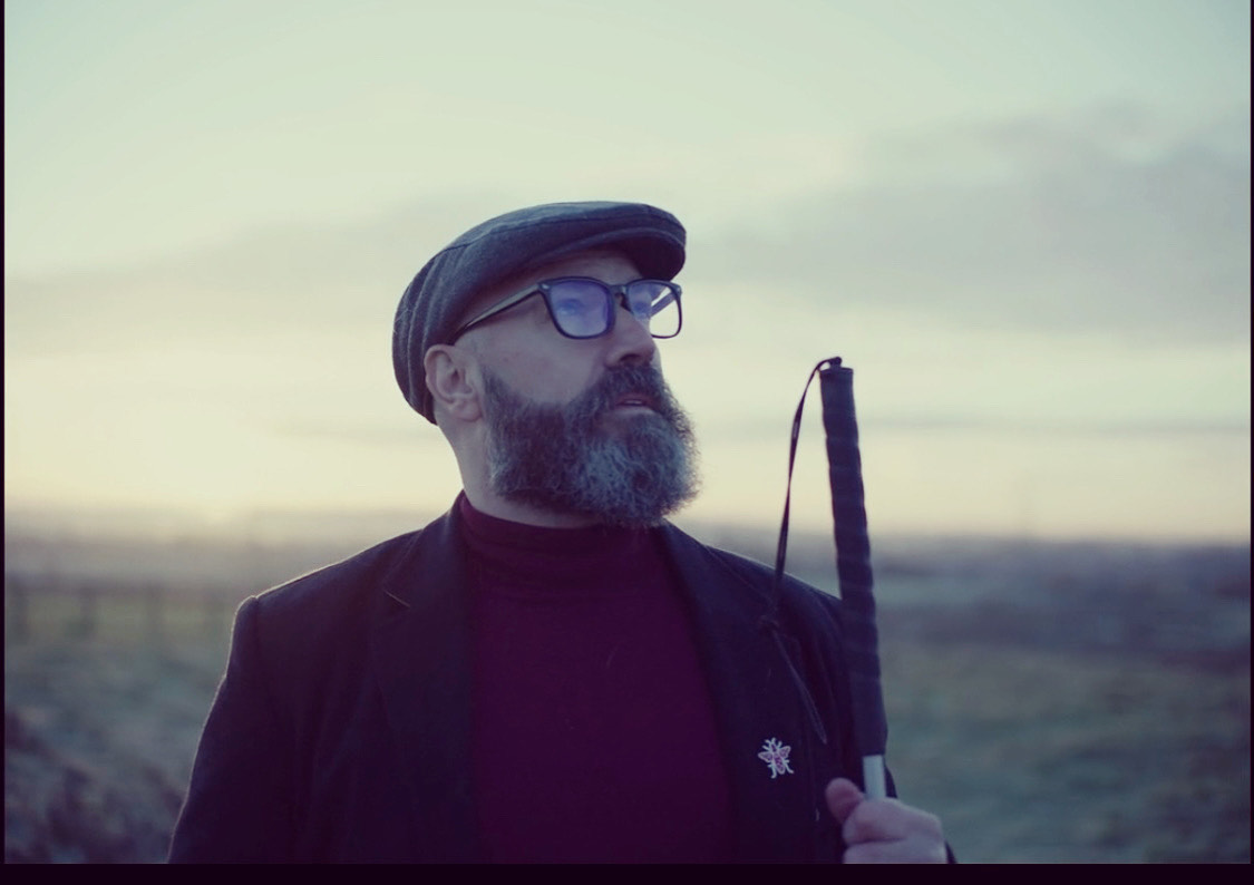 Dave Steele standing outdoors with hazy background of a field and cloudy sky. Dave is holding up his cane. He is wearing a grey cap, glasses, dark blazer with a motif on the lapel and a maroon turtle neck sweater.