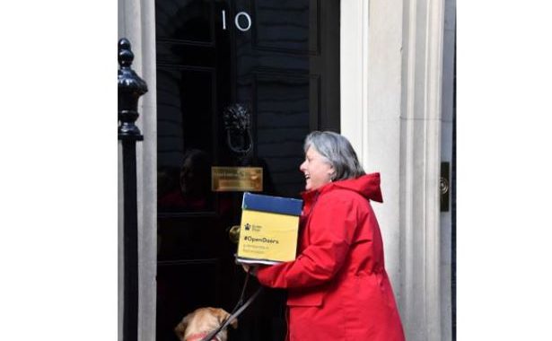 Campaigner Penny in a red jacket with her yellow lab guide dog handing in a box to someone inside No 10 Downing Street.