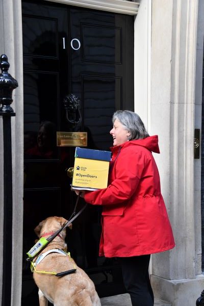 Campaigner Penny in a red jacket with her yellow lab guide dog handing in a box to someone inside No 10 Downing Street.