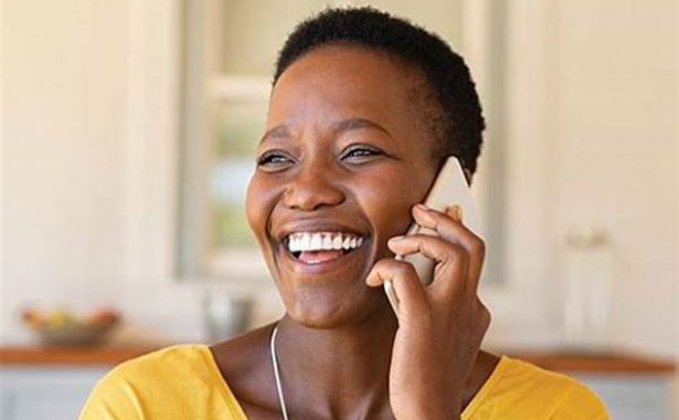 A Afro-Caribbean woman speaking on a phone. She is smiling an wearing a yellow top.