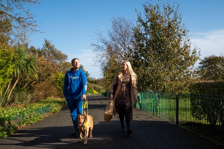 A vision impaired person with their guide dog and a member of the Vision Support Barrow & District team walking side by side through Barrow Park.