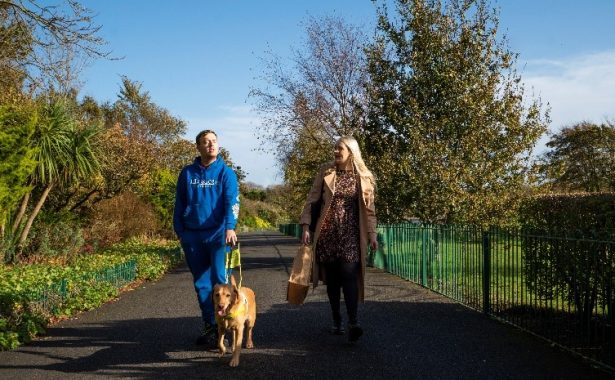 A vision impaired person with their guide dog and a member of the Vision Support Barrow & District team walking side by side through Barrow Park.