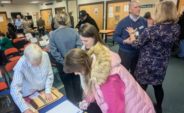 A room full of men and women of various ages talking to each other. At the front is Christine with a woman and her daughter looking at a display on a table. Behind them Jacquie is speaking to Trustee Campbell Chalmers,